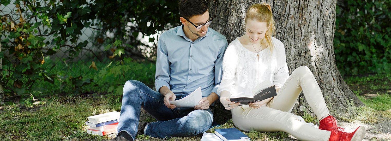 Theologie studieren an der Katholischen Privat-Universität Linz. Ein männlicher und eine weibliche Studierende sitzen im Freien an einen Baumstamm gelehnt und lesen in Büchern.