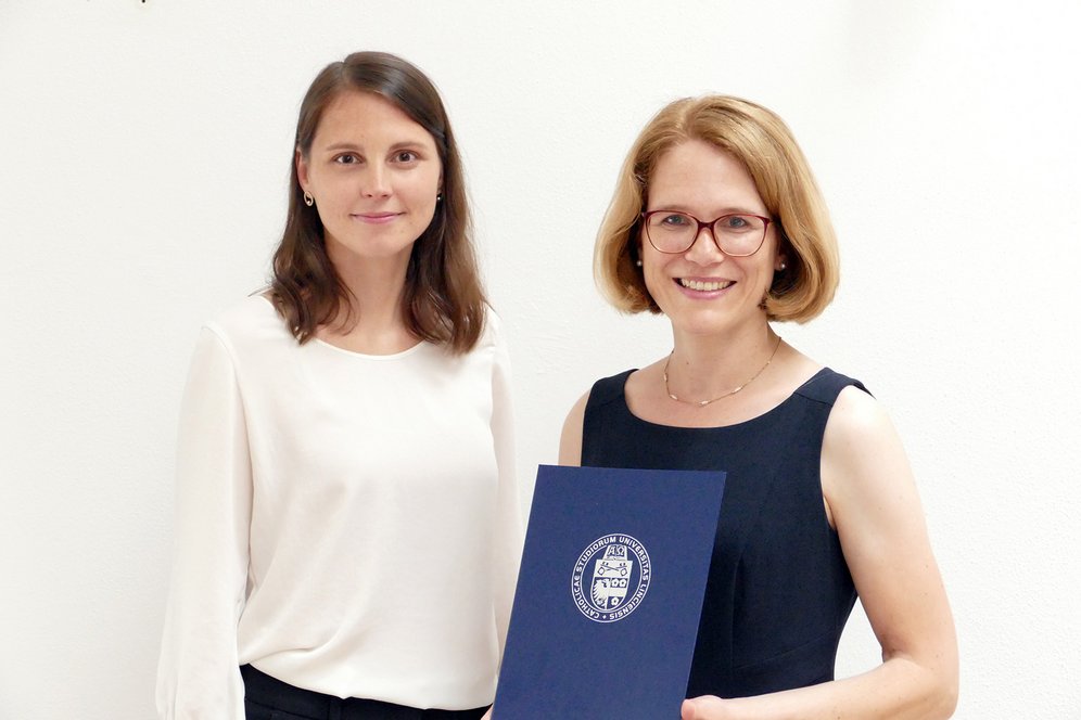 Links steht eine brünette Frau, rechts steht eine blonde Frau und hält eine Urkunde.